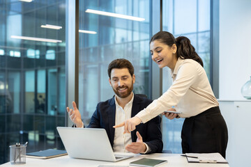 A businesswoman and businessman are collaborating on a laptop in a modern office, sharing a moment of discussion and a smile.