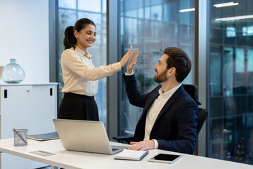 A diverse pair of colleagues celebrates success with a high-five in a modern office setting. Sunlight streams through the windows.