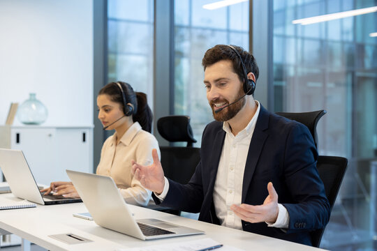 A man in a suit enthusiastically gestures during a video call, while a woman works at a computer in the background. Both wear headsets.