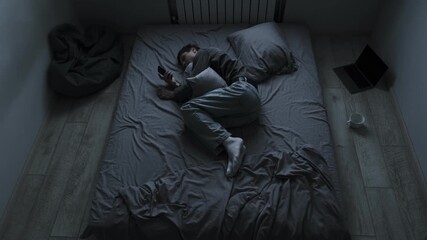 Broken heart after breakup. Young man lying in bed hugging pillow looking at photos of ex girlfriend feeling pain and wanting to text her. - Powered by Adobe