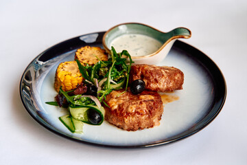 A beautifully plated serving of meat patties accompanied by grilled corn, fresh greens, and a creamy dipping sauce, highlighting a delicious culinary experience.