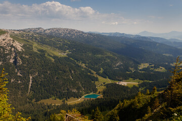 Naklejka premium Panoramic view from Walmendingerhorn, showcasing lush green valleys, dense pine forests, and a vibrant turquoise mountain lake under a blue sky.