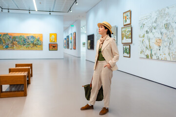 Museum day. Wide shot of young Caucasian pretty woman in elegant suit and straw hat posing in the art gallery. Concept of cultural education