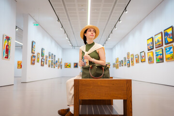 Wide shot of elegant young Caucasian woman in straw hat sitting in the art gallery. Lots of paintings in the background. Concept of cultural education and Museum Day