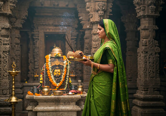 South Asian Woman in Green Silk Saree Carrying Puja Thali to Lord Shiva's Shrine in Intricately Carved Temple