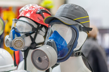 Indoor scene featuring two mannequins, one with a red helmet and goggles, the other in a gray cap Both wear white respirators A third figure partially visible in background wears a yellow helmet A