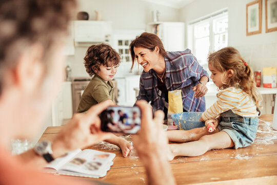 Happy young family taking a photo with a smartphone while being messy and baking with flour at home in the kitchen - Powered by Adobe