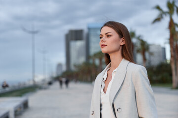 Business woman in light coat walking outdoors in city with serious confident expression and modern buildings background