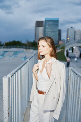 Fototapeta premium Young woman in stylish white suit walking in urban environment during cloudy day with modern buildings in background, fashion and business concept