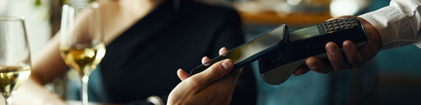 Caucasian young adult woman making contactless payment with smartphone while holding wine glass, male hand holding wireless card reader, restaurant setting visible in background