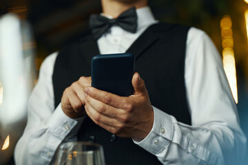 Caucasian young adult man wearing formal attire using smartphone while standing indoors, hands and torso visible, focusing on device, not looking at camera, background blurred