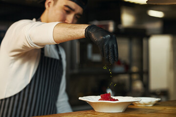 Young adult Asian man wearing black gloves garnishing dish with herbs in professional kitchen, focusing on precise plating technique, preparing food for restaurant service
