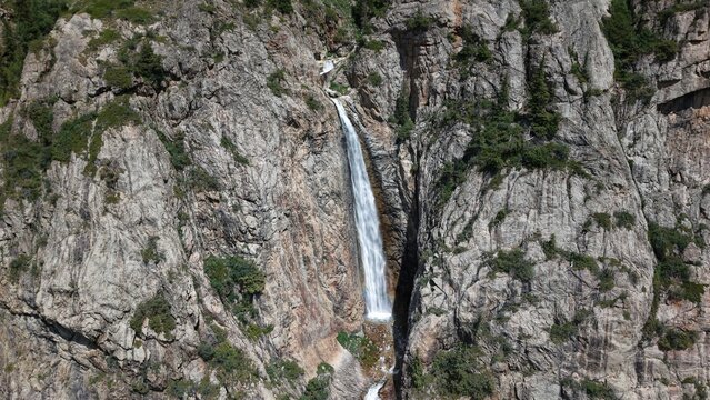 Refreshing waterfall cascading down a steep, rocky mountainside, surrounded by lush greenery, creating a picturesque natural landscape on a sunny summer day