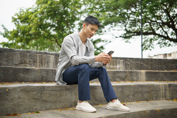 Obraz premium Young asian man sitting on outdoor stone steps, using his smartphone in a peaceful park environment