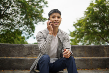 Smiling male in casual outfit talking on smartphone while relaxing on stone steps outside.
