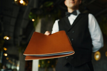 Caucasian young adult man wearing formal vest and bow tie handing menu to customer in restaurant, focus on menu held in foreground with server slightly out of focus in background