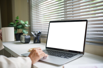 Fototapeta premium Close up of businesswoman working on laptop with a blank white screen, holding a coffee mug in a bright office.