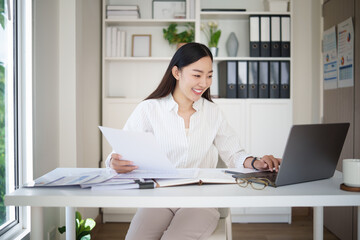 Smiling businesswoman working on laptop while reviewing financial reports at modern office desk.