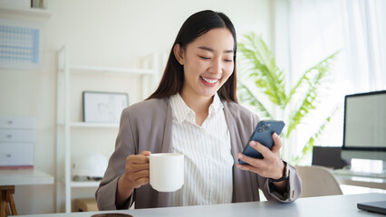 Smiling businesswoman enjoying coffee while checking smartphone in modern office.