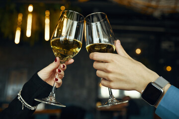 Caucasian woman and Caucasian man clinking wine glasses during celebration, closeup of hands with jewelry and smartwatch, blurred background suggesting indoor social gathering
