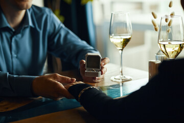 Caucasian young adult man proposing marriage by presenting engagement ring to young adult woman at restaurant table, both holding hands, wine glasses visible in foreground