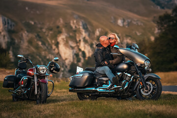 Happy couple kissing on a black cruiser motorcycle in scenic mountain landscape, enjoying a romantic sunset moment during a motorbike road trip.