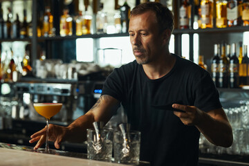Middle aged Caucasian man preparing cocktail behind bar counter, holding black saucer and arranging glassware, liquor bottles displayed on shelves in background
