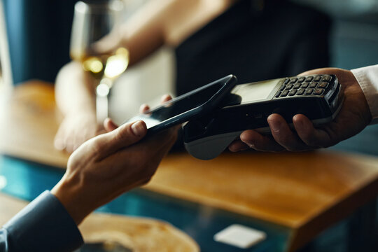 Caucasian young adult man holding smartphone making contactless payment using wireless terminal while sitting at table in restaurant, female figure with wine glass visible in background