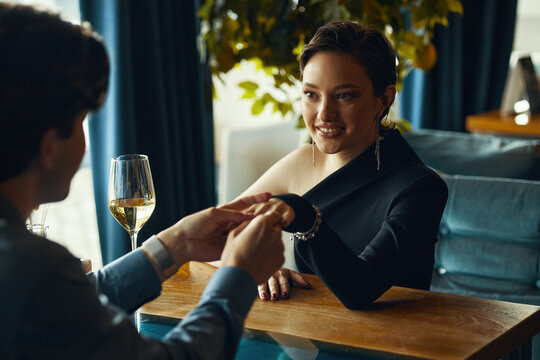 Caucasian young adult woman smiling while holding hands with Caucasian young adult man across table in restaurant, wine glass on table, woman making eye contact with man - Powered by Adobe