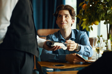 Young adult Caucasian man sitting at restaurant table handing credit card to waiter holding payment terminal, smiling and making eye contact, wine glasses and tree in background