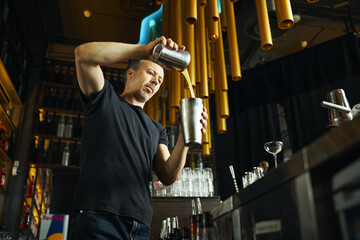 Caucasian middle aged man preparing cocktail behind bar counter, pouring liquid from shaker into mixing tin, surrounded by bar equipment and glassware, focused on bartending task