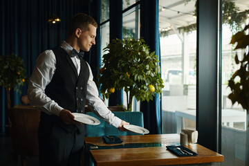 Caucasian young adult man wearing formal vest and bow tie setting table in restaurant, holding...