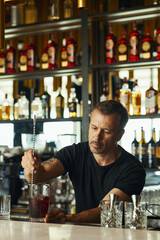 Middle aged Caucasian man stirring cocktail in mixing glass behind bar counter, concentrating on preparing drink with bottles of alcohol arranged on shelves in background