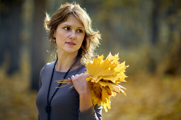 A beautiful young woman with windswept hair holds a vibrant bouquet of golden maple leaves, looking thoughtfully to the side in a soft-focused autumn park background.