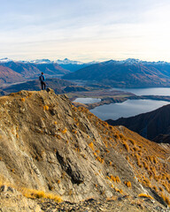 Hiker man stands on summit of Roys Peak and admires stunning panoramic view over Lake Wanaka and surrounding snow covered peaks near Wanaka, Otago, South Island, New Zealand