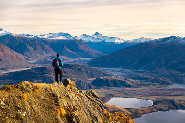 Hiker man stands on summit of Roys Peak and admires stunning panoramic view over Lake Wanaka and surrounding snow covered peaks near Wanaka, Otago, South Island, New Zealand