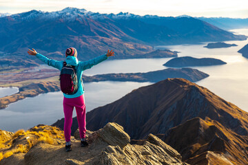 Hiker woman stands on summit of Roys Peak with arms outstretched admires breathtaking panoramic view over Lake Wanaka and surrounding snow covered peaks near Wanaka, Otago, South Island, New Zealand