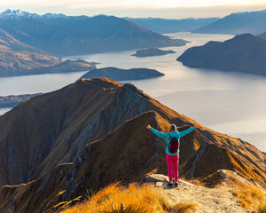 Hiker woman stands on summit of Roys Peak with arms outstretched and admires breathtaking panoramic view over Lake Wanaka and surrounding snow covered peaks near Wanaka, Otago, South Island, New Zeala