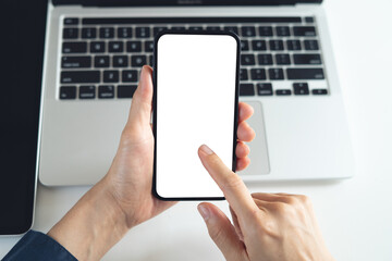 Blank white screen cell phone mockup. Close up, business person using smartphone, finger touching on screen during working on laptop computer on office table, for social media marketing, overhead shot