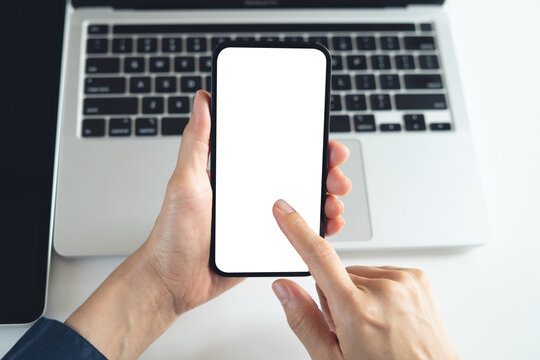 Blank white screen cell phone mockup. Close up, business person using smartphone, finger touching on screen during working on laptop computer on office table, for social media marketing, overhead shot