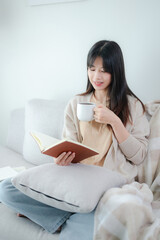 A young Asian woman is comfortably nestled on a sofa at home, engrossed in reading a book while...