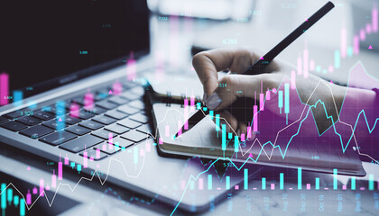 Financial trader writing in notebook with digital forex and stock market graphs overlayed on laptop background in modern office environment.