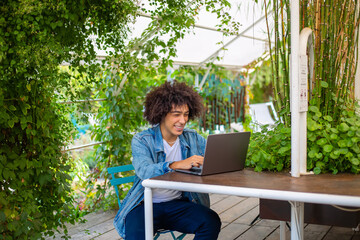Smiling young ethnic guy  old, dressed in casual clothes, works on a laptop outdoors, in a green area in nature. Green open space for coworking, freelancer, relaxing, coffee, food. Free place to work