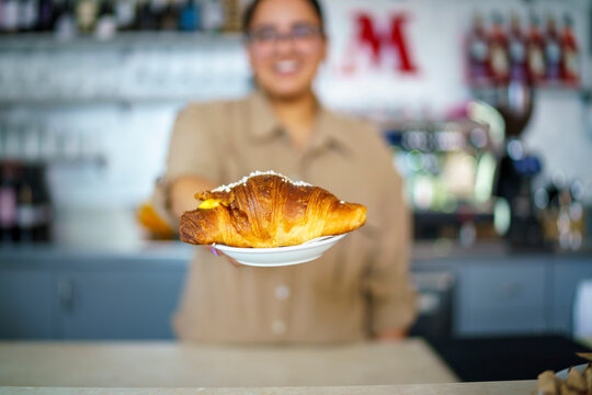 Friendly smiling barista holding a delicious golden brown croissant topped with white sprinkles on a white plate at a cozy cafe counter interior background with blurred coffee machines