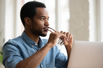 Portrait of thoughtful african american man working on laptop with glasses in mouth