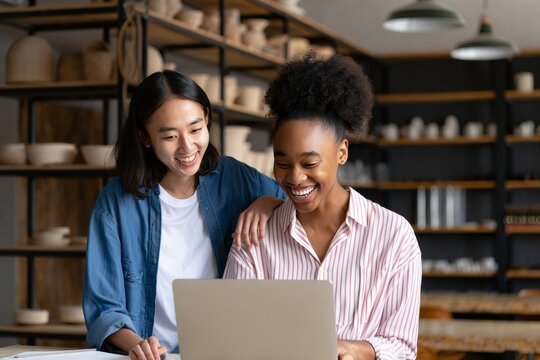 Two women working together on a laptop in a pottery studio smiling and collaborating