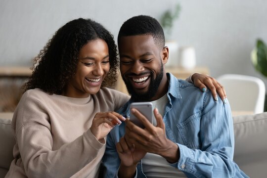 Happy african american couple laughing looking at smartphone at home together relax