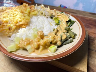 pecel is a stew of long beans, sprouts and mustard greens sprinkled with crushed peanut sauce and peanut brittle as a complement, a close up of warm pecel rice served on a ceramic plate on the table