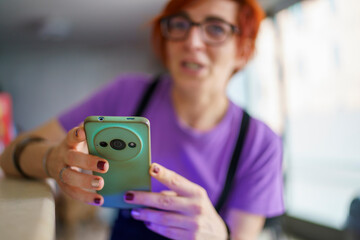 A middle-aged woman with short red hair and glasses holding a smartphone and speaking during a casual outdoor gathering creating a vibrant and engaging scene