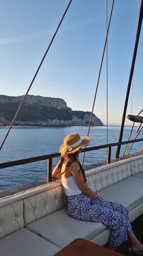 Relaxed young woman tourist wearing a straw hat, sailing in a voyage boat, looking out at the sea and mountains in the background under a sunset sky. Beautiful Turkish Mediterranean coastline of Kemer